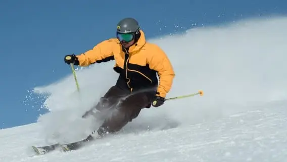 Man skiing with a big plume of snow behing wearing yellow and black ski suit and a dark grey helmet