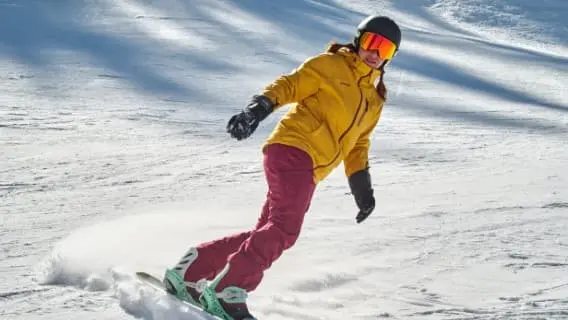 Female snowboarder making a gentle turn, wearing yellow jacket, pink trousers and a reflective visor