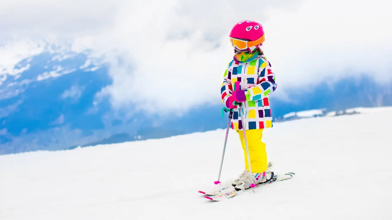 small cbhild wering a multicoloured ski jacket standing on the ski slope with tiny skis