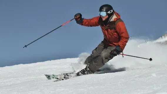 Man in red ski jacket, grey trousers and black helmet skiing fast with spray of snow